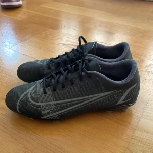 Nike Mercurial svarta fotbollsskor - Svarta Nike Mercurial fotbollsskor med grå detaljer och snörning. Skorna har en smal, sportig siluett och mönstrad ovandel i syntetmaterial. Utrustade med fasta dobbar för gräsplan. Perfekta för dig som vill ha stilrena och snabba skor på planen.