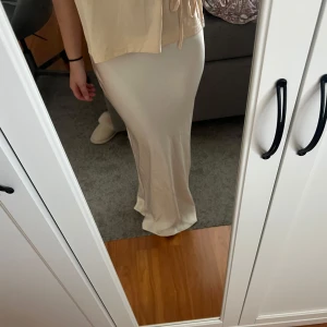 Vit långkjol i satin från Zara - Säljer en elegant vit långkjol från Zara i mjukt satinmaterial. Kjolen har en slät och glansig yta med en figurnära passform som faller snyggt längs benen. Perfekt för dig som gillar stilrena och minimalistiska plagg.