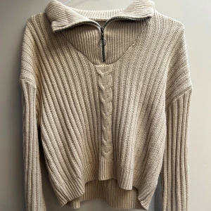 Beige stickad tröja med half zip - Beige stickad tröja med ribbat mönster och snygg kabelstickad detalj framtill. Tröjan har en hög krage med half zip och långa ärmar. Perfekt för lager på lager och chill stil under höst och vinter.