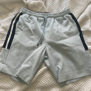 Ljusgrå shorts från BOSS med snörning - Snygga ljusgrå shorts från BOSS med svarta och vita ränder längs sidorna. De har elastisk midja med snörning och är perfekta för en avslappnad stil. Mjukt material som känns skönt mot huden.