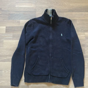 Mörkblå stickad kofta från Polo Ralph Lauren - Snygg mörkblå stickad kofta från Polo Ralph Lauren med dragkedja och ribbad hög krage med ljusgrå detalj. Klassisk broderad logga på bröstet och två fickor framtill. Perfekt för lager-på-lager och chill stil.