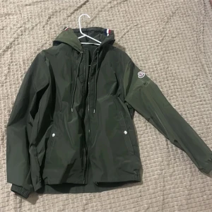 Grön Moncler vindjacka med huva - Snygg grön vindjacka från Moncler med huva och dragsko. Jackan har Moncler-logga på ärmen, dragkedja framtill och praktiska fickor med knappar. Perfekt för dig som vill ha en stilren och sportig look. Passar till både vår och höst.
