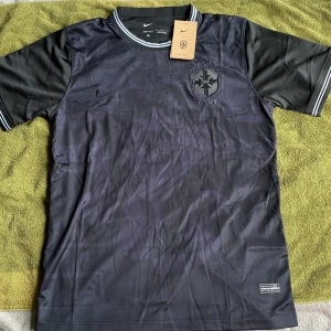 Svart Brasilien Nike fotbollströja - Snygg svart Brasilien fotbollströja från Nike med Dri-FIT-material. Tröjan har korta ärmar, diskret camouflagemönster och broderat CBF-märke på bröstet. Ribbad krage och ärmslut med gråa detaljer. Perfekt för dig som vill sticka ut på planen.