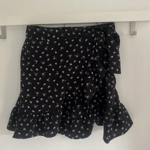 Svart blommig omlottkjol från FB Sister - Supersöt svart omlottkjol från FB Sister med små vita blommor och volangkant. Kjolen har knyt i sidan och är perfekt för dig som gillar en feminin och trendig stil. Kort modell som passar till många olika looks.