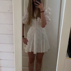 Vit blommig kortklänning från Adoore - Supersöt vit kortklänning från Adoore med broderade blommor, puffiga trekvartsärmar och djup v-ringning. Perfekt för sommarens alla tillfällen! Endast använd en gång så är i perfekt skick! Prefekt som studentklänning eller konfirmationsklänning!!☀️🌸