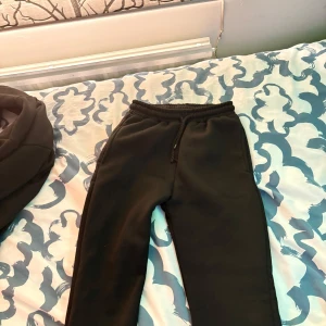 Central cee syna world set  - Svarta mjukisbyxor med grön broderad text 'Forgive Me' på ena benet. Byxorna har resår och snörning i midjan för skön passform. Perfekta för chill dagar eller streetwear-stil.