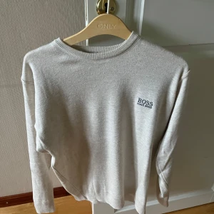 Beige tröja från Hugo Boss - Snygg beige tröja från Hugo Boss med diskret logga på bröstet. Tröjan har rund halsringning och långa ärmar, perfekt för en clean och stilren look. Mjuk och skön, passar till det mesta i garderoben.