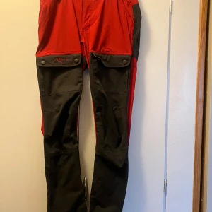 Röda och svarta friluftsbyxor High Mountain - Riktigt snygga röda och svarta friluftsbyxor från High Mountain. Byxorna är tillverkade i en mix av polyester, bomull och elastan för extra komfort och rörelsefrihet. Perfekta för utomhusaktiviteter och äventyr. Storleken motsvarar ungefär 38-40 dam men med korta ben. Stretchiga lår men åt det större hållet i midjan. Något blekta längst ner i bensluten. Hela och rena! Noppor kan förekomma.
