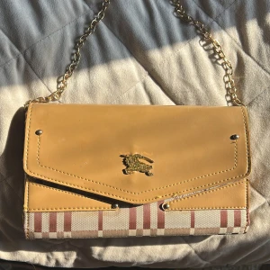 Beige kuvertväska med kedja Burberry - Använd kuvertväska från Burberry i beige och brunt rutmönster, med guldfärgad kedja och detaljer. Väskan har klassisk Burberry-logga framtill, flera fack och kortplatser inuti samt dragkedja. Material i skinn och textil, perfekt för att bära det viktigaste. Lite sliten därav priset. 
