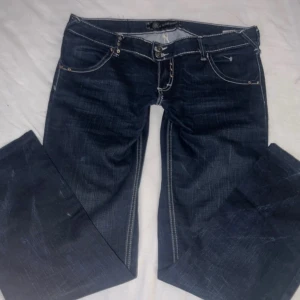 Ultra low waits jeansbyxor med vita sömmar från Rock Revival - Säljer ett par mörkblå jeansbyxor från Rock Revival med tydliga vita kontrastsömmar och dekorativa detaljer på bakfickorna. Byxorna har normal passform, låg midja och knappar framtill. Snygga detaljer med nitar och broderi på fickorna.