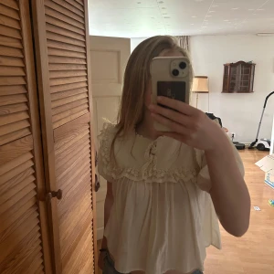 Söt volangblus från Zara i offwhite - Romantisk blus från Zara i offwhite med stickad överdel, volangkant och knytning vid halsen. Puffiga korta ärmar och fina spetsdetaljer ger en vintagekänsla. Perfekt för dig som gillar en gullig och trendig stil. Har en fläck som eventuellt går bort i tvätten. Kan tvättas och dubbelkollas innan köp.