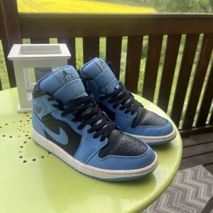 Nike Air Jordan 1 Mid blå och svarta sneakers - Säljer ett par Nike Air Jordan 1 Mid i blått och svart med vita detaljer. Skorna har klassisk snörning, rund tå och platt sula. Perfekta för dig som gillar streetwear och vill sticka ut med en ikonisk modell.