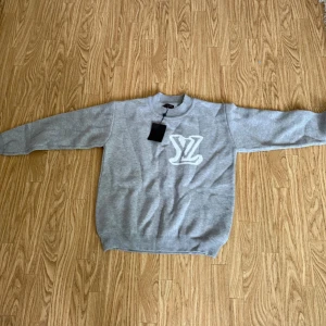 Grå stickad tröja med LV-logga - Säljer en grå stickad tröja från Louis Vuitton med vit LV-logga både fram och bak. Tröjan har rund halsringning och långa ärmar. Perfekt för dig som gillar stilrena och exklusiva plagg.