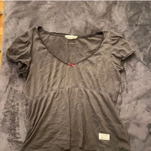 Grå ribbad topp med rosett - Supersöt grå ribbad topp från Odd Molly med korta ärmar och en liten rosa rosett vid den djupa v-ringningen. Toppen har en lätt utsvängd passform och en liten etikett nertill. Perfekt för dig som gillar detaljer och en feminin stil.