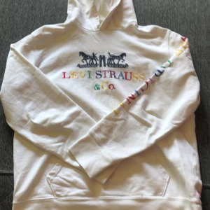 Vit hoodie från Levi's med regnbågsbrodyr - Vit hoodie från Levi's i bomull med broderad logga och regnbågsfärgade bokstäver på bröstet och ena ärmen. Klassisk känguruficka och huva. Perfekt för dig som gillar färgglada detaljer och streetstyle.