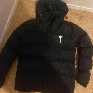 Svart pufferjacka från Trapstar - Säljer en svart pufferjacka från Trapstar med huva och dragkedja. Jackan har logga på bröstet och stor Trapstar-tryck på ryggen. Perfekt för kalla dagar och riktigt snygg streetstil.