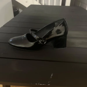 Svart lackad Mary Jane-klack - Säljer en svart, glansig Mary Jane-sko med bred klack och rund tå. Snygg rem med knappdetalj över foten. Perfekt för dig som vill ha en klassisk och stilren sko med lite retrovibbar.