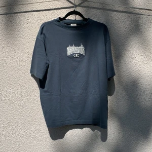 Blå t-shirt från Champion med tryck - Säljer en blå t-shirt från Champion i 100% bomull. Framsidan har ett broderat Champion-tryck med stadssilhuett. Oversized passform och korta ärmar. Storlek S. Knappt använd.
