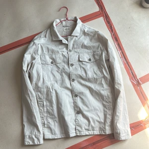 Beige overshirt från East West - Säljer en stilren beige overshirt från East West i bomull. Jackan har klassisk krage, knäppning framtill, två bröstfickor med lock och knappar samt knappar vid ärmsluten. Perfekt för lager-på-lager och enkel att matcha.