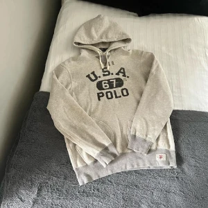 Grå hoodie från Polo Ralph Lauren - Snygg grå hoodie från Polo Ralph Lauren med tryck 'U.S.A. 67 POLO' på bröstet. Klassisk känguruficka, vita snören i huvan och ribbade muddar. Perfekt för chill dagar och streetstyle. Mjuk bomullskänsla och relaxed fit. Passar S bra med