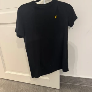 Svart t-shirt från Lyle & Scott - Svart t-shirt från Lyle & Scott med klassisk passform och rund hals. T-shirten har det ikoniska gula fågel-loggan broderad på bröstet. Tillverkad i mjuk bomull och har korta ärmar. Perfekt basic-plagg med stilren look.
