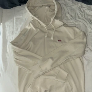 Beige hoodie från Levi's med huva - Snygg beige hoodie från Levi's med klassisk huva och snören. Tröjan har en liten röd Levi's-logga på bröstet. Tröjan har aldrig används då den va lite stor så den är i helt nytt skick.