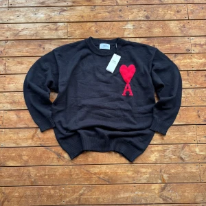Ami Paris Sweater - Svart stickad tröja från Ami med rund halsringning. På bröstet finns en röd broderad hjärtformad detalj med bokstaven A. Tröjan har långa ärmar och ribbade muddar vid ärmslut och nederkant. Perfekt för en stilren och trendig look. Pris är diskuterbart.
