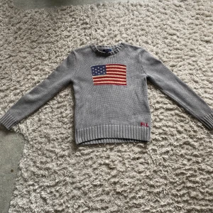 Ralph Lauren tröja  - En trendig grå Ralph lauren tröja med en amerikansk flagga som tryck. Storlek 14-16 viket motsvarar XS. Den är använd men funkar som den ska dock tyvärr en liten grön fläck på tröjans framsida. Hör av er vid minsta intresse då priset går att ändras på!