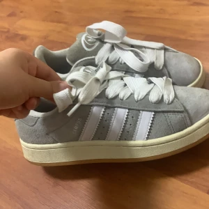 Adidas campus  - Säljer ett par grå Adidas Campus sneakers med klassiska vita ränder och vit sula. Skorna har breda vita skosnören och är i mocka med Adidas-logga bak. Perfekta för en stilren och avslappnad look.