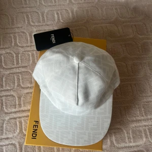Vit keps från Fendi - Vit keps från Fendi med diskret mönster av Fendi-loggor i tyget. Klassisk form med böjd skärm och justerbar passform. Kepsen är tillverkad i bomull och har en exklusiv känsla, perfekt för en stilren look.