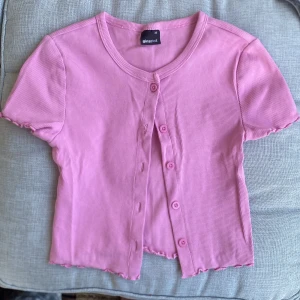 Rosa ribbad kortärmad topp från Gina Tricot - Säljer en söt rosa ribbad topp från Gina Tricot med korta ärmar och knappar framtill. Toppen har vågiga kanter vid ärmslut och nederkant för en extra detalj. Perfekt att styla till jeans eller kjol!