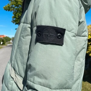Ljusgrön pufferjacka från Stone Island - Säljer en ljusgrön pufferjacka från Stone Island med svart logotyp-patch på ärmen. Även en färgad logotyp-patch på insidan som man kan byta till. Jackan har dragkedja framtill, två fickor och huva. Perfekt för kyliga dagar och riktigt snygg streetstil.