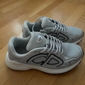 Gråa sneakers från Dior - Säljer ett par gråa Dior sneakers med tydlig CD-logga på sidan. Skorna har vit och svart sula, mesh- och syntetdetaljer samt snörning. Perfekta för dig som gillar stilrena och exklusiva sneakers. Fick den i födelsedagspresent o använt en gång. Pris kan diskuteras vid snabbaffär!