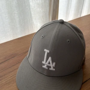 Grå LA-keps från New Era - Säljer en stilren grå keps från New Era med LA-logga broderad framtill i vitt. Klassisk modell med böjd skärm och ventilerande hål. Perfekt för dig som gillar sportig streetstyle.