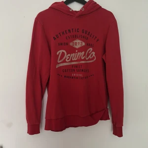 Röd hoodie från Produkt Denim Co - Röd hoodie från Produkt Denim Co med tryck framtill. Tröjan har huva med dragsko och långa ärmar. Perfekt för en avslappnad stil och enkel att matcha med jeans eller joggers.