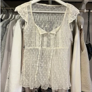 Vit spetsblus med prickigt mönster - Ej mina bilder! Säljer en söt och romantisk vit blus i transparent spets med små prickar och fina spetsdetaljer vid axlarna. Blusen har v-ringning, knytband framtill och är ärmlös. Perfekt för lager-på-lager eller till sommarens outfits.
