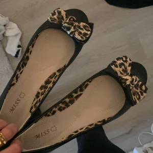 Svarta ballerinaskor med leopardrosett från miss Miss - Säljer ett par svarta ballerinaskor med öppen tå och leopardmönstrad rosett framtill. Insidan har också leoparddetaljer som ger skorna en extra touch. Perfekta för dig som vill sticka ut lite extra! Lägre pris kan diskuteras🩷