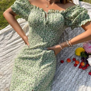 Grön blommig klänning - Säljer en söt grön kortklänning med blommigt mönster. Klänningen har volangdetaljer vid ärmarna och en liten knytning framtill. Perfekt för varma dagar och somriga tillfällen💕