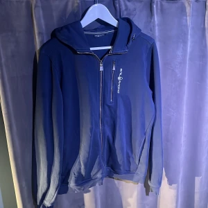 Marinblå hoodie från Sail Racing - Säljer en marinblå hoodie från Sail Racing med dragkedja framtill och logga på bröstet. Tröjan har huva och en bröstficka med dragkedja. Perfekt för svala dagar och enkel att matcha med det mesta.