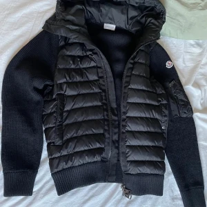 Svart Moncler dunjacka med stickade ärmar - Snygg svart dunjacka från Moncler med stickade ärmar och huva. Jackan har quiltad front, dragkedja och Moncler-logga på ärmen. Perfekt för dig som vill ha en stilren och varm jacka med exklusiv känsla.