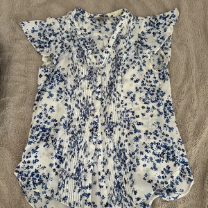 Vit blommig blus - Supersöt vit blus från H&M med blått blommönster och korta, volangiga ärmar. Blusen har knappar framtill och fina veckdetaljer längs bröstet. Perfekt för en fräsch och somrig look!