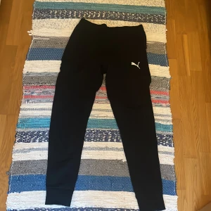 Svarta mjukisbyxor från Puma - Svarta mjukisbyxor från Puma med klassisk logga på låret. Byxorna har en slim passform med muddar vid bensluten och är tillverkade i mjukt material, perfekta för chill eller träning.