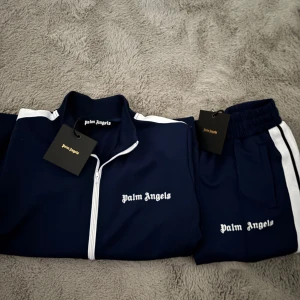 Mörkblå Palm Angels dress - Säljer en mörkblå dress av märket Palm Angels med vita ränder längs ärmarna och vit dragkedja. Jackan har hög krage och logga broderad på bröstet. Perfekt för en sportig & stilig look.