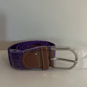 Lila flätat bälte  - Säljer ett snyggt lila flätat bälte med detaljer i brunt läder och ett klassiskt silverfärgat spänne. Perfekt accessoar för att ge outfiten lite extra färg. Bältet har vita sömmar på läderdetaljerna och är flexibelt i materialet.