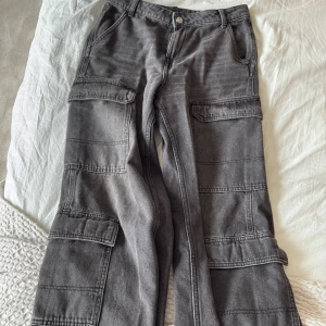 Grå cargopants från H&M Divided - Säljer ett par grå cargopants från H&M Divided med bred passform och flera stora fickor på benen. Byxorna är tillverkade i jeansmaterial och har en cool, avslappnad vibe. Perfekta för dig som gillar streetstyle och vill ha något bekvämt men ändå trendigt.