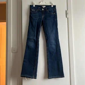 Blå bootcut jeans med fickdetaljer - Snygga mörkblå jeans med bootcut-modell och klassiska kontrastsömmar. Byxorna har låg midja och dekorativa knappar på bakfickorna. Har sprättat upp längs ned på bena. Kontakta mig vid frågor eller funderingar😊                        Midjemått: 38cm                                                       Innerbenslängd: 79cm