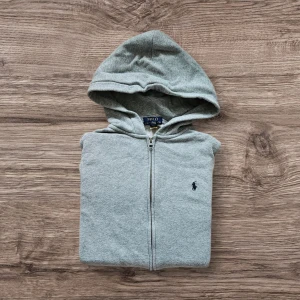Ralph Lauren Zip hoodie - Säljer en Grå Zip hoodie från Ralph Lauren eftersom den inte används. Tröjan har krympt lite i tvättmaskinen men annars inga fläckar eller liknande. Hör gärna av er vid frågor, priset är ej hugget i sten. 
