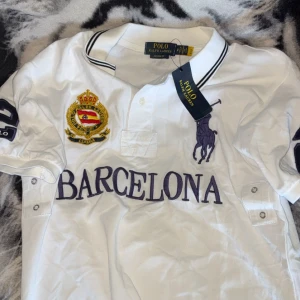 Vit piké från Polo Ralph Lauren med Barcelona-tryck - Vit pikétröja från Polo Ralph Lauren med stor lila logga, broderad patch och texten BARCELONA på framsidan. FETT tröja mycket bra kvalitet.
