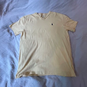 Gul t-shirt från Polo Ralph Lauren - Snygg gul t-shirt från Polo Ralph Lauren med klassisk v-ringning och liten broderad logga på bröstet. Tillverkad i 100% bomull, perfekt för en chill och stilren look. Enkel design som funkar till det mesta. Aa fett till sommaren