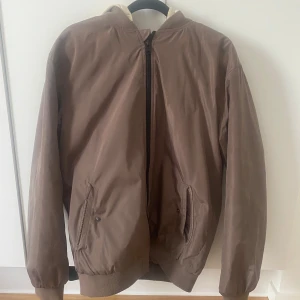 Beige bomberjacka med avtagbar huva - Snygg beige bomberjacka från Kappahl med avtagbar ljus huva. Jackan har ribbade muddar, dragkedja framtill och två fickor med tryckknapp. Perfekt för dig som gillar en clean och stilren look med streetvibe.